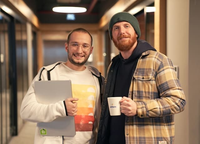 Dois profissionais sorrindo em um corredor de escritório moderno, um segurando um notebook e o outro uma caneca de café.