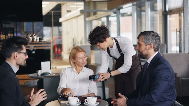 Grupo de pessoas em reunião em uma cafeteria enquanto um atendente passa o pagamento na maquininha.