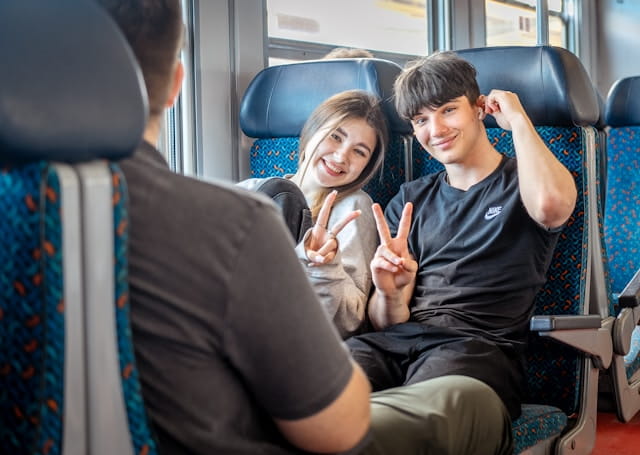 Dois estudantes sorrindo dentro de um trem fazendo sinal de paz para a câmera.